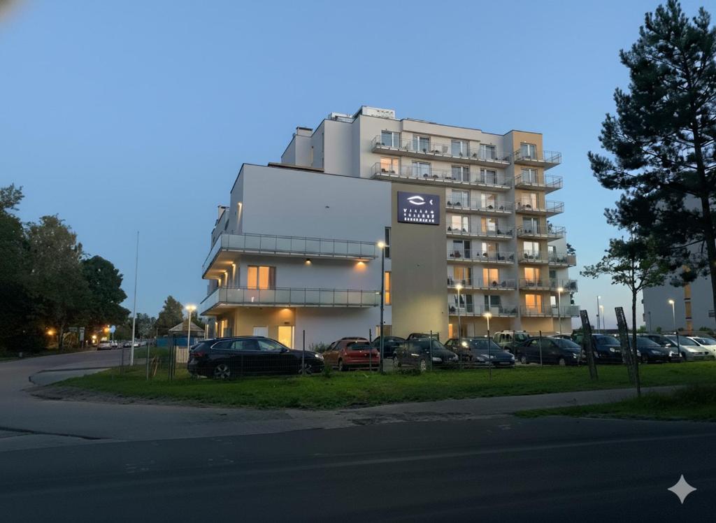 Modern multi-story waterfront hotel with lit balconies and parked cars, calm twilight sky.