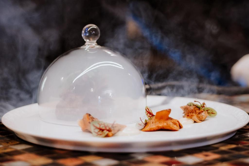 Glass cloche releasing smoke over small dumplings and garnishes on white plate, dramatic presentation.