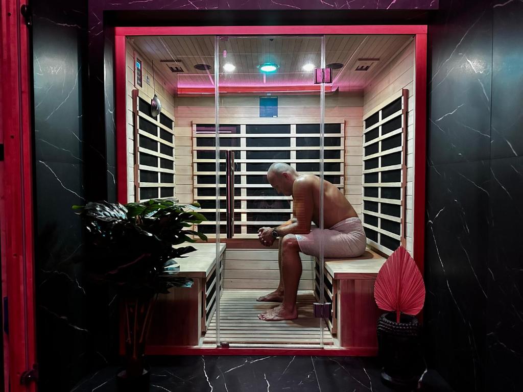 Man wrapped in towel seated inside a small glass-front sauna with wooden benches and plants.