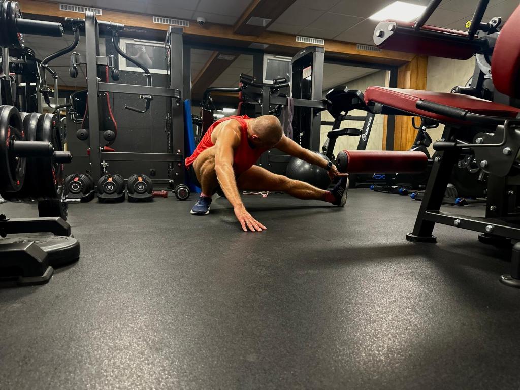 Person in red tank top performing seated leg stretch on gym floor amid weights.