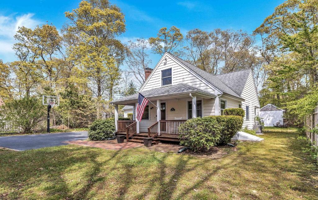 a white house with a porch and an american flag at Jenkins Pond Beach Access with BBall Hoop & Grill, Bright Cape Cod Retreat near Jenkins Pond in Falmouth - Parking, Modern Retreat with Outdoor Space, 24 Hour Hospitality in Falmouth