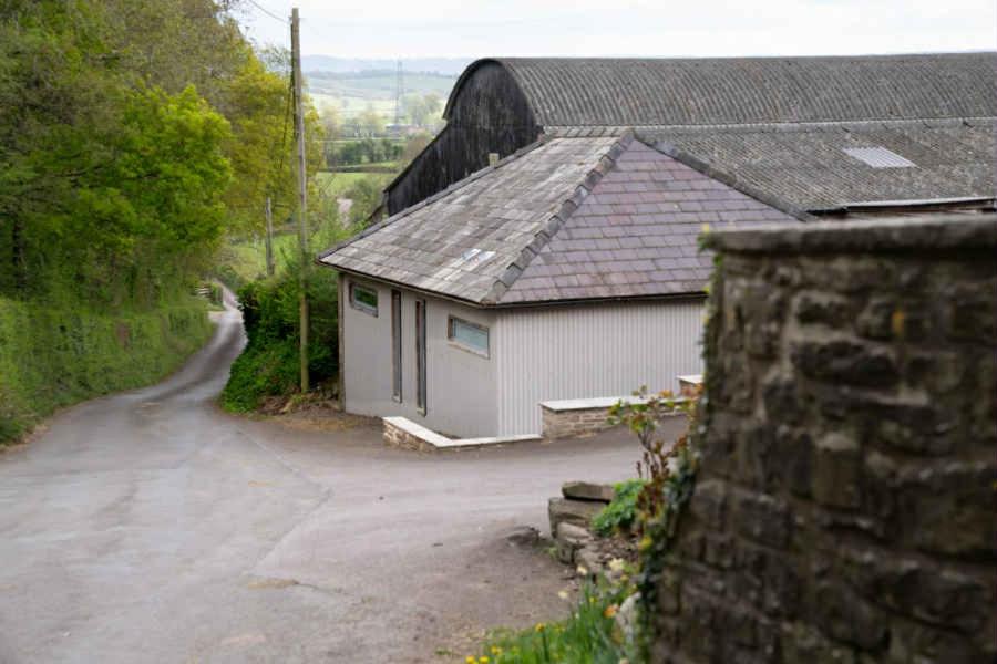 a small white building with a roof next to a road at The Cartshed in Llanvetherine