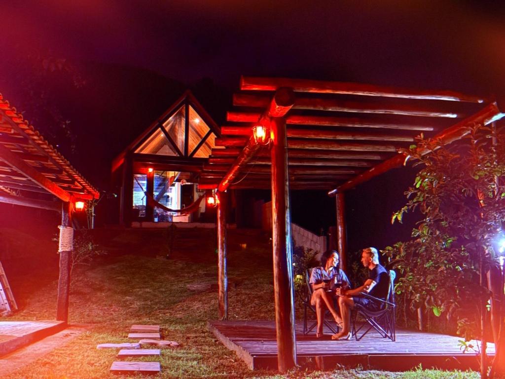 two people sitting in chairs under a pergola at night at Chalé Sertão da Boa Vista in Rio Claro