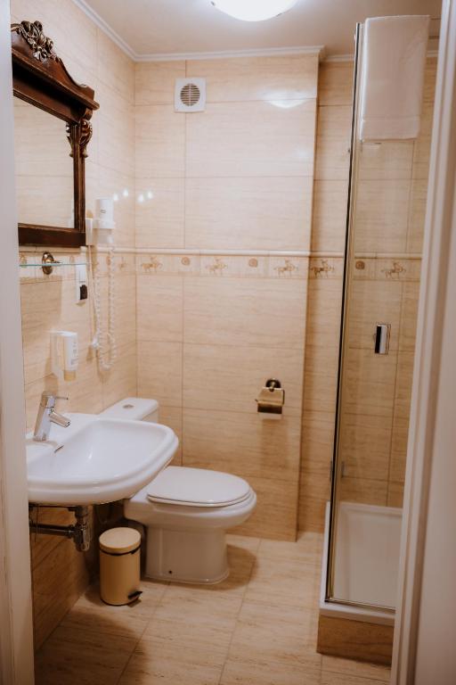 Compact beige-tiled bathroom with pedestal sink, toilet, ornate framed mirror, and glass shower enclosure.