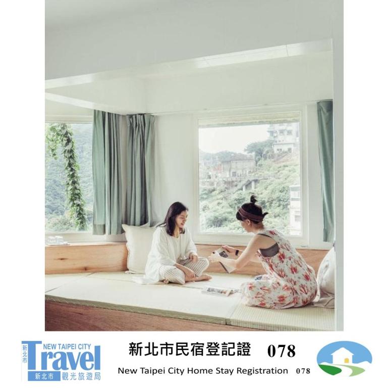 Zwei Frauen sitzen auf einem Bett vor einem Fenster in der Unterkunft 九份綠光 故事所 山中夢遊 - OwlStay Jiufen Green Ray in Jiufen
