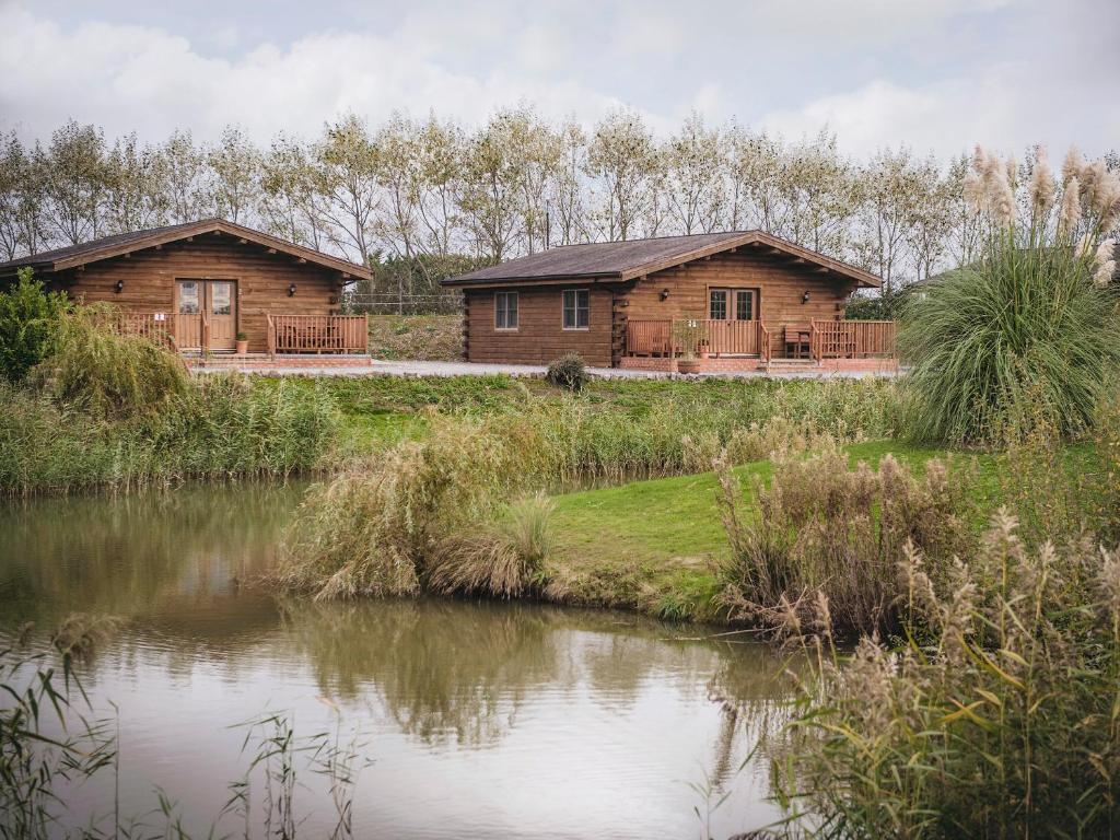 two wooden huts next to a river with trees at Lake View Lodges - 25076 in Old Leake