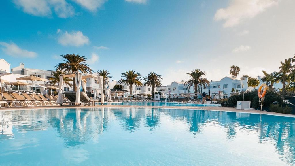 a large swimming pool with palm trees and white buildings at BLUESEA Corralejo Village in Corralejo