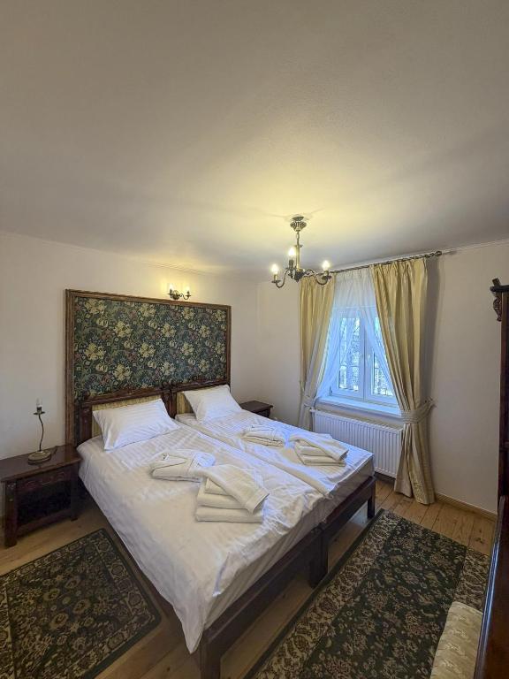 Bright bedroom with double bed, floral headboard, folded towels, window draped with cream curtains.