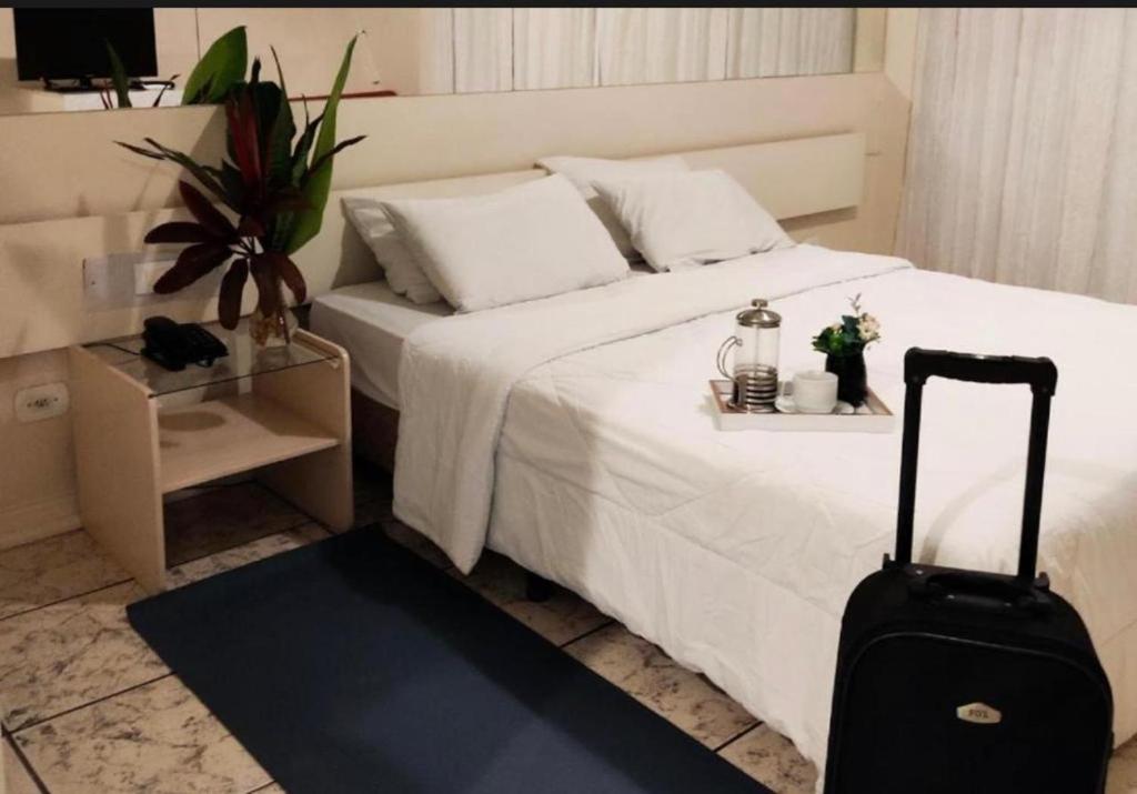 a bedroom with a white bed with a suitcase at Quarto Hotel Palhano londrina in Londrina