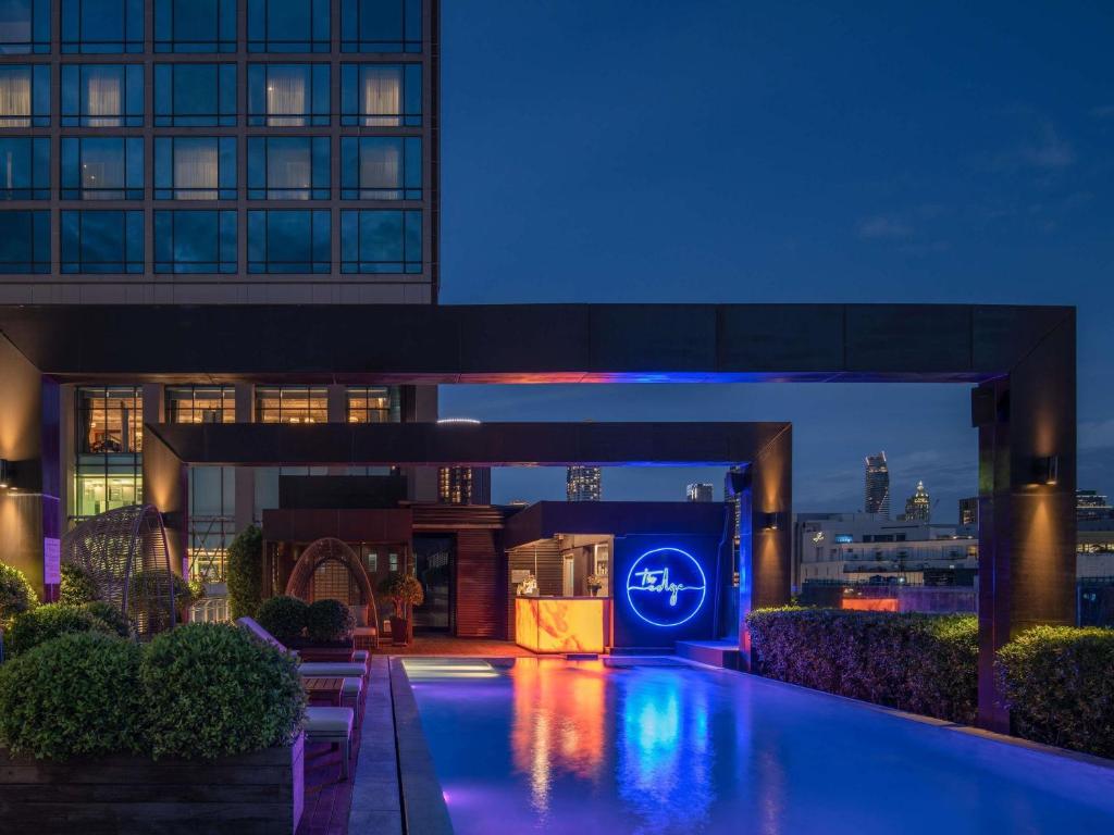 A rooftop pool glows at night beneath the hotel and a Bangkok skyline.