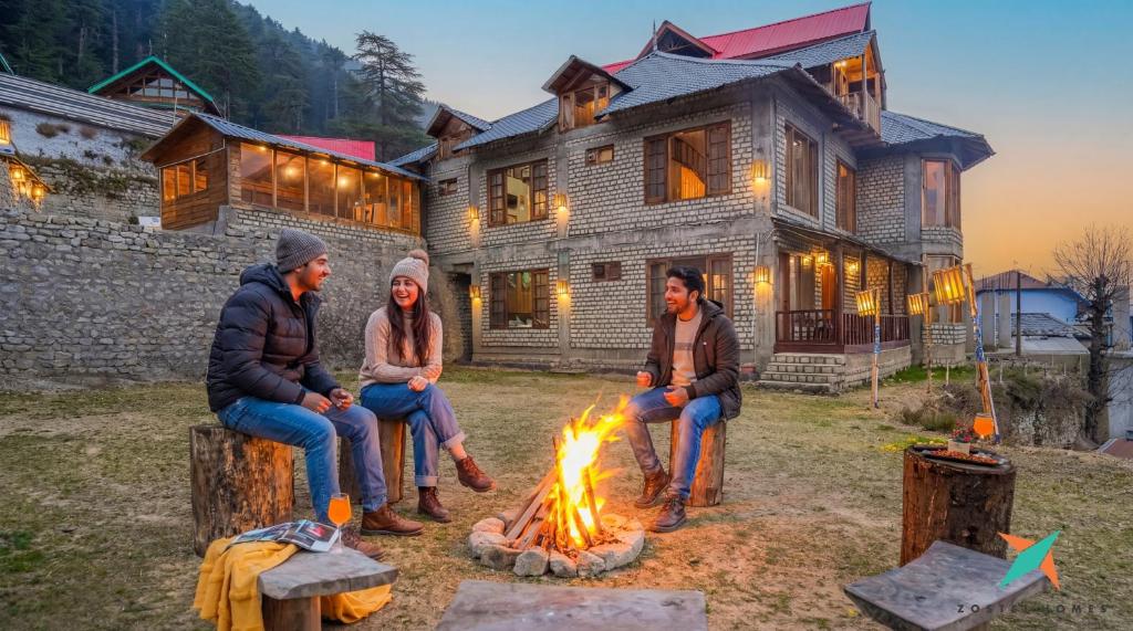 three people sitting around a fire in front of a house at Zostel Homes Khajjiar in Chamba