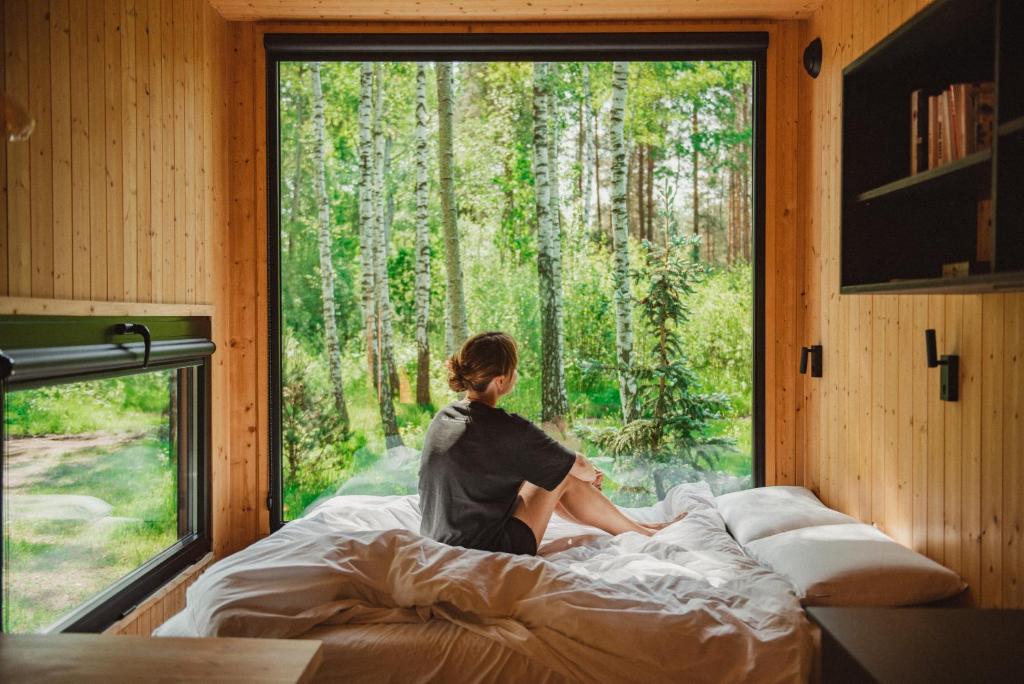 a woman sitting on a bed looking out a window at Hidden Base - romantyczna leśna kryjówka in Smolarnia
