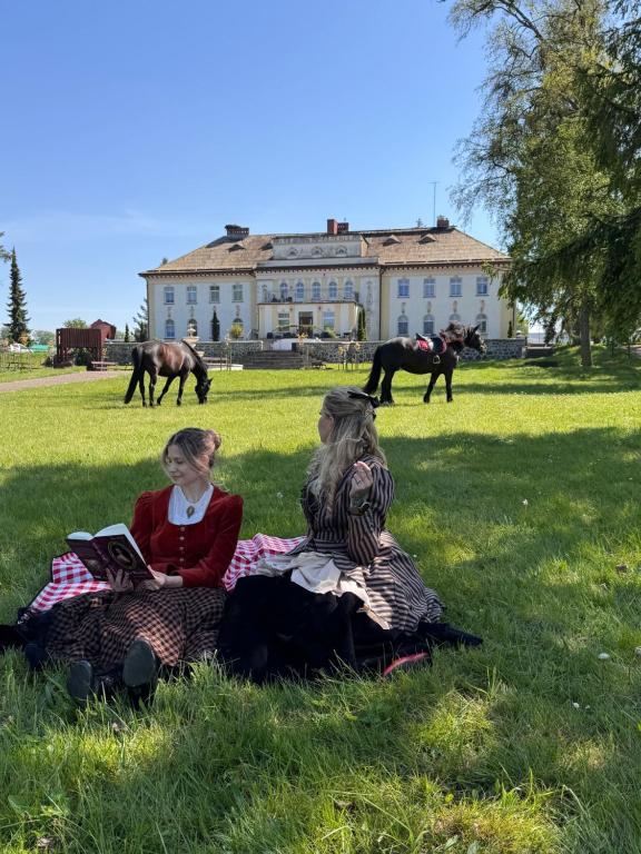 two women sitting on the grass in a field with horses at Pałac pod Bocianim Gniazdem in Runowo