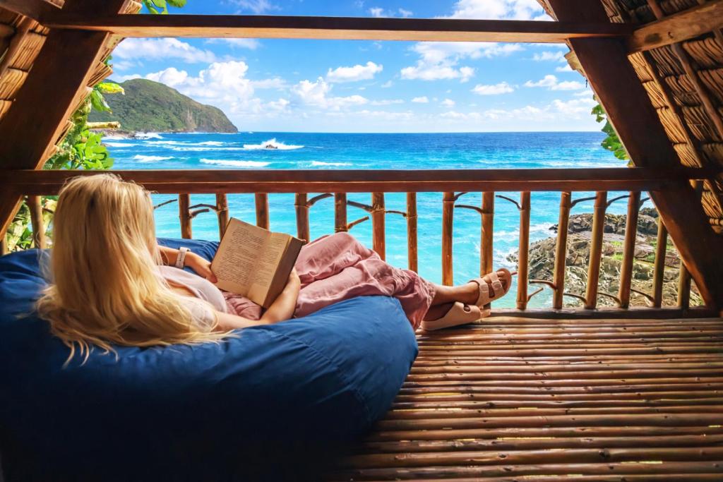 a woman sitting on a couch reading a book on a balcony overlooking the ocean at L'Astrolabe - Private Beach in Baras