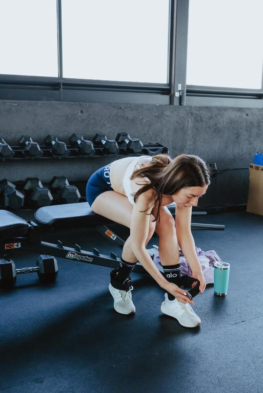 A woman ties her shoes beside free weights and a workout bench.