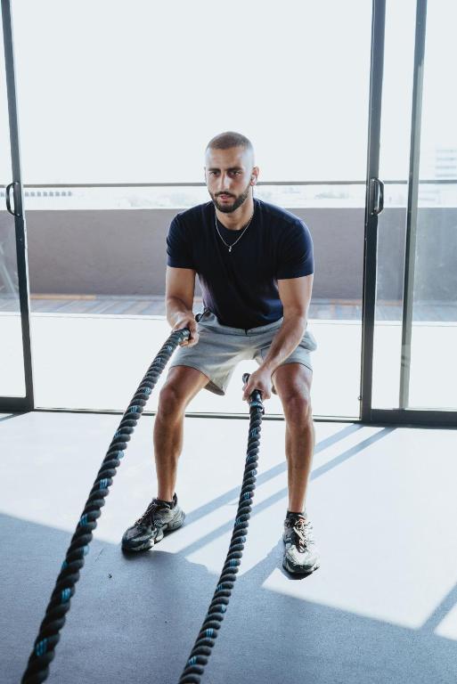 A man uses battle ropes in a bright gym beside floor-to-ceiling windows.