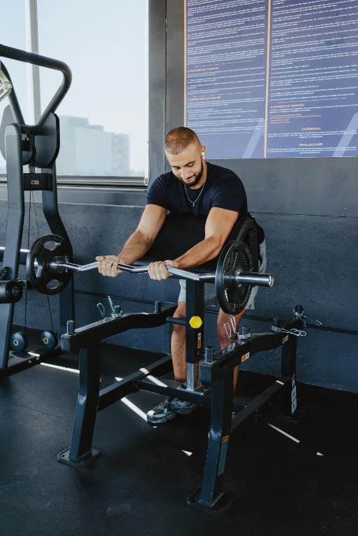 A man performs a barbell exercise on a bench in a windowed gym.