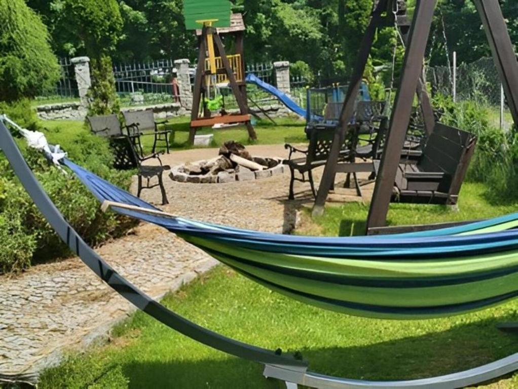 Striped green-blue hammock stretched across sunny garden with cobblestone path, benches, and central fire pit.