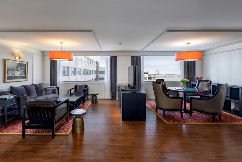 An expansive hotel living area with sofas, lounge chairs, a dining table and river-facing windows.