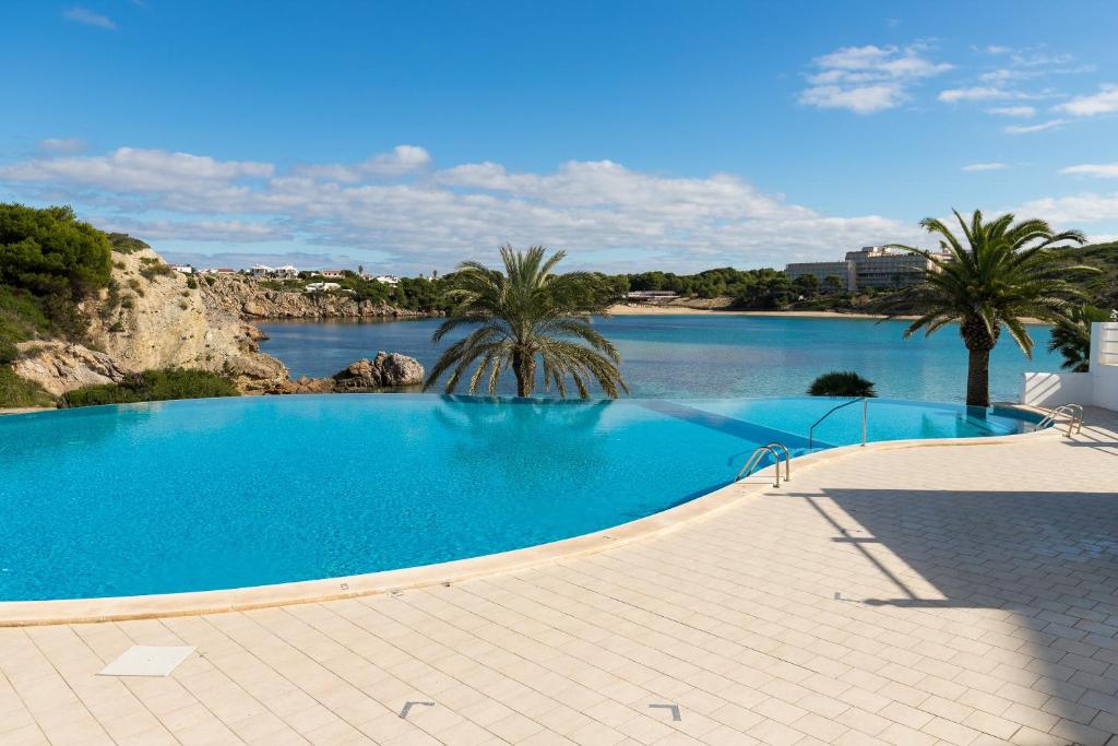 a large swimming pool with palm trees and a body of water at White Sands Maluz in Arenal d'en Castell