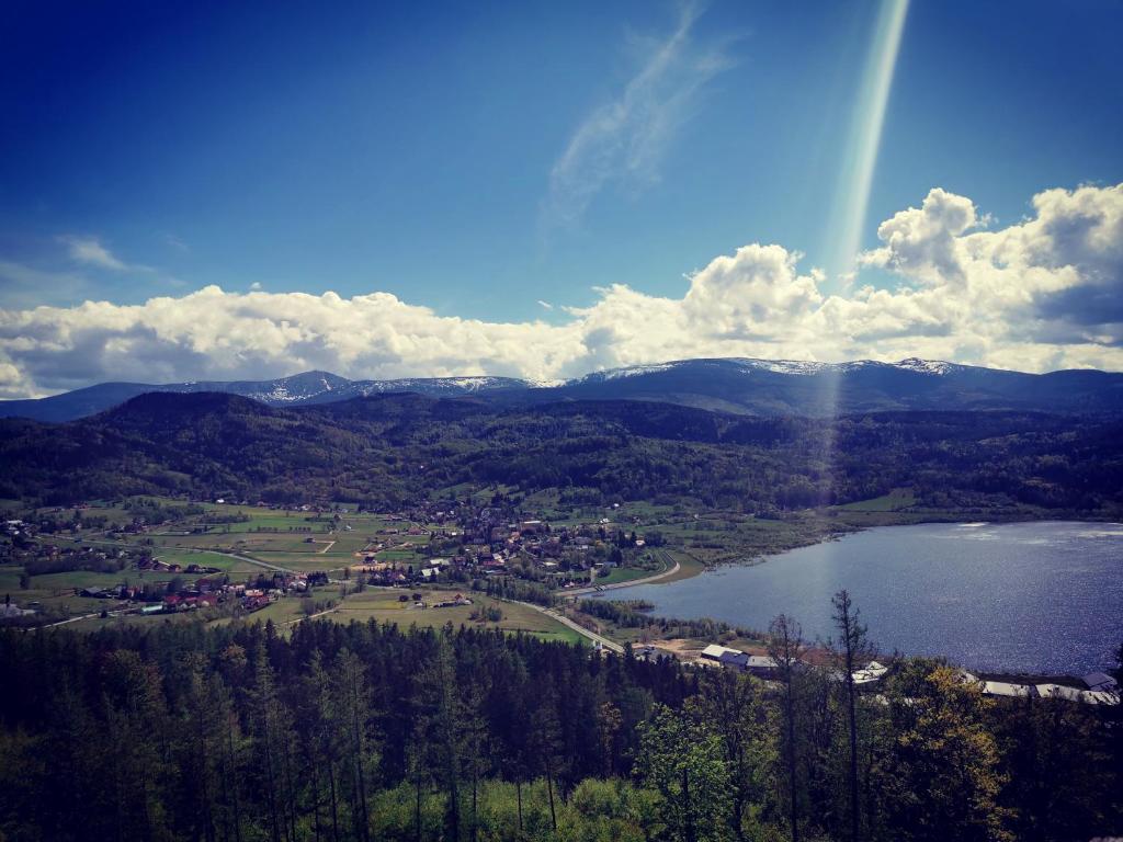 Panoramic valley with village, lake and distant snow-capped mountains under a sunlit sky.