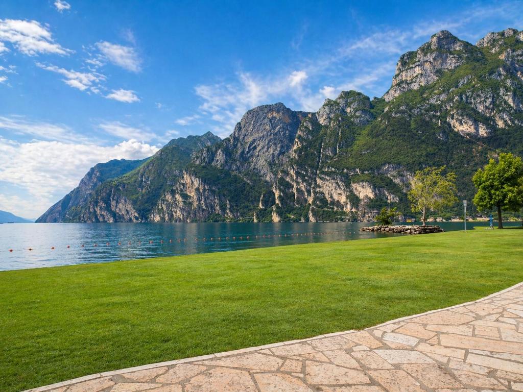 a grassy field next to a body of water with mountains at Oxygen Riva del Garda Monolocale con Balcone e Bike Room in Riva del Garda