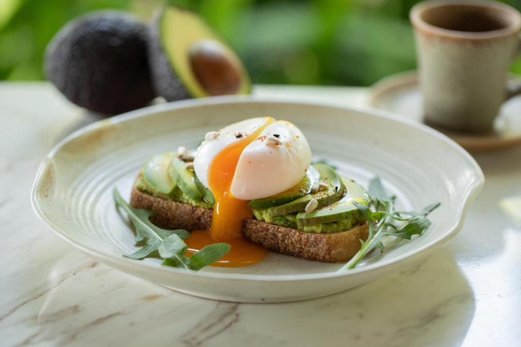 Avocado toast with a soft egg is presented on a bright marble table beside coffee.