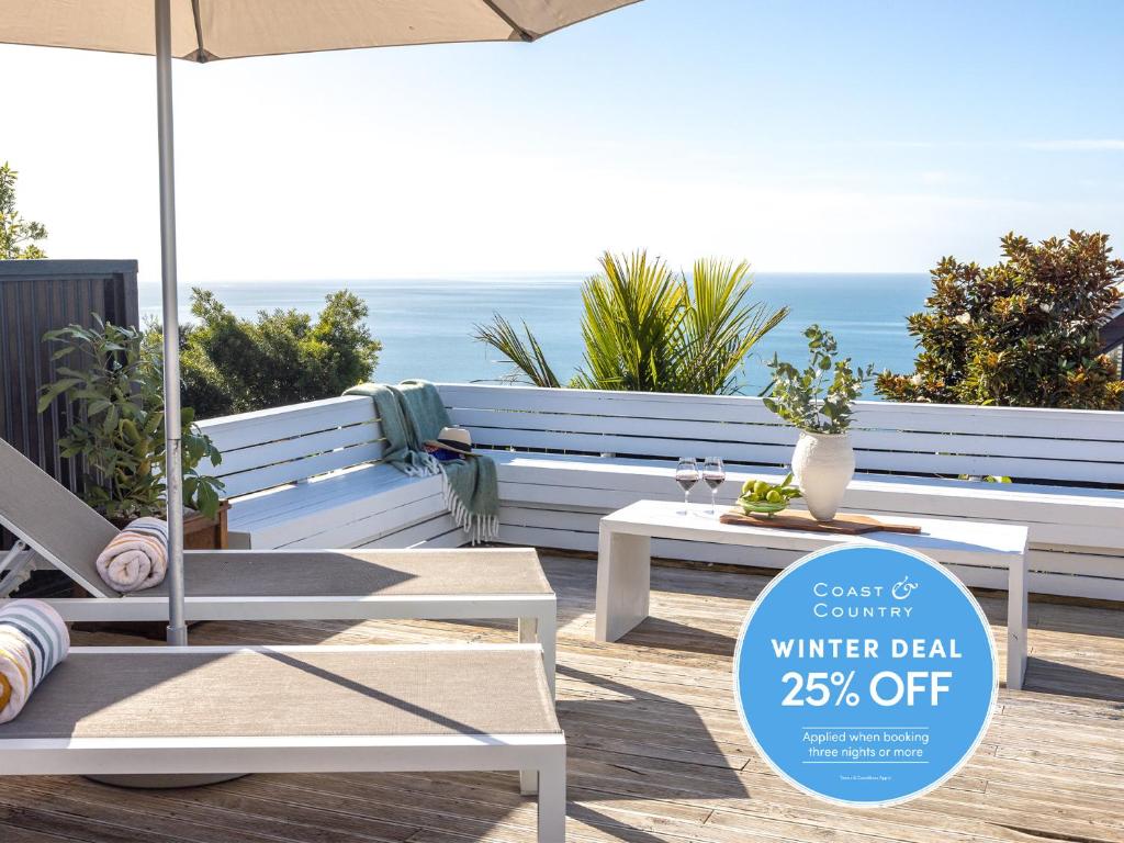 a patio with white benches and an umbrella and the ocean at Ocean Breeze Cottage - Coast & Country in Onetangi