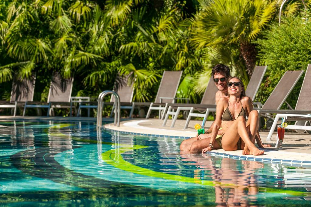 Ein Mann und eine Frau sitzen auf einem Surfbrett im Pool. in der Unterkunft Parc Hotel Flora in Riva del Garda