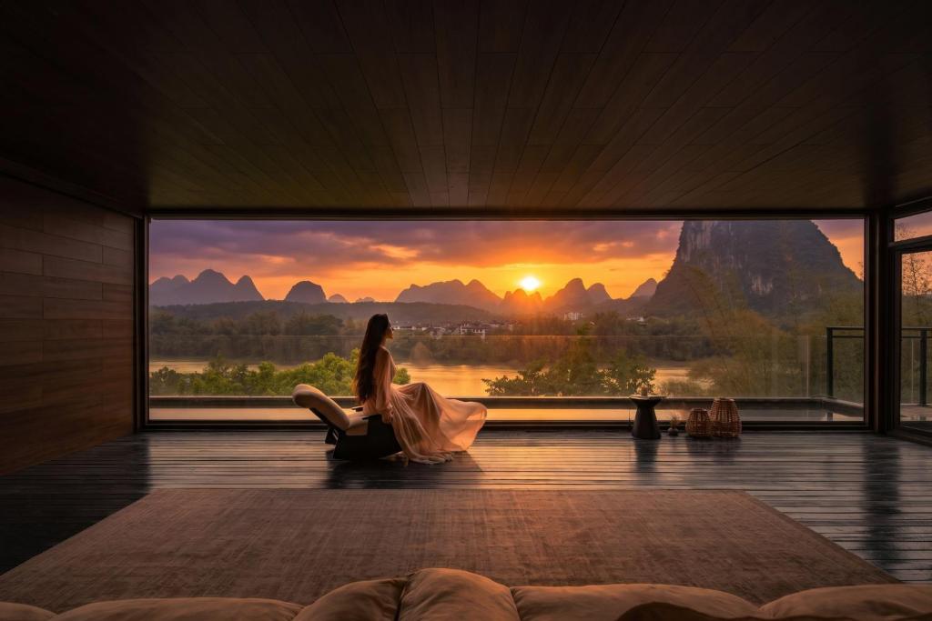 a woman sitting in a room looking out a window at a sunset at Yangshuo Gallery Resort--River View, Sunset,Cafe,Karast Mount,Ten-Mile Gallery,Free Laundry,Travel Guide,Book Tickets in Advance,E-Scooter Service in Yangshuo