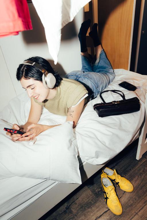 a woman wearing headphones laying on a bed with a cell phone at Hostel by Hyve Basel in Basel