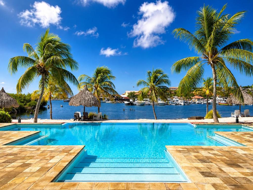 a swimming pool with palm trees and a body of water at JeVakantie Spanish Water Apartments B7 and C8 with Infinity Pool near Jan Thiel in Willemstad
