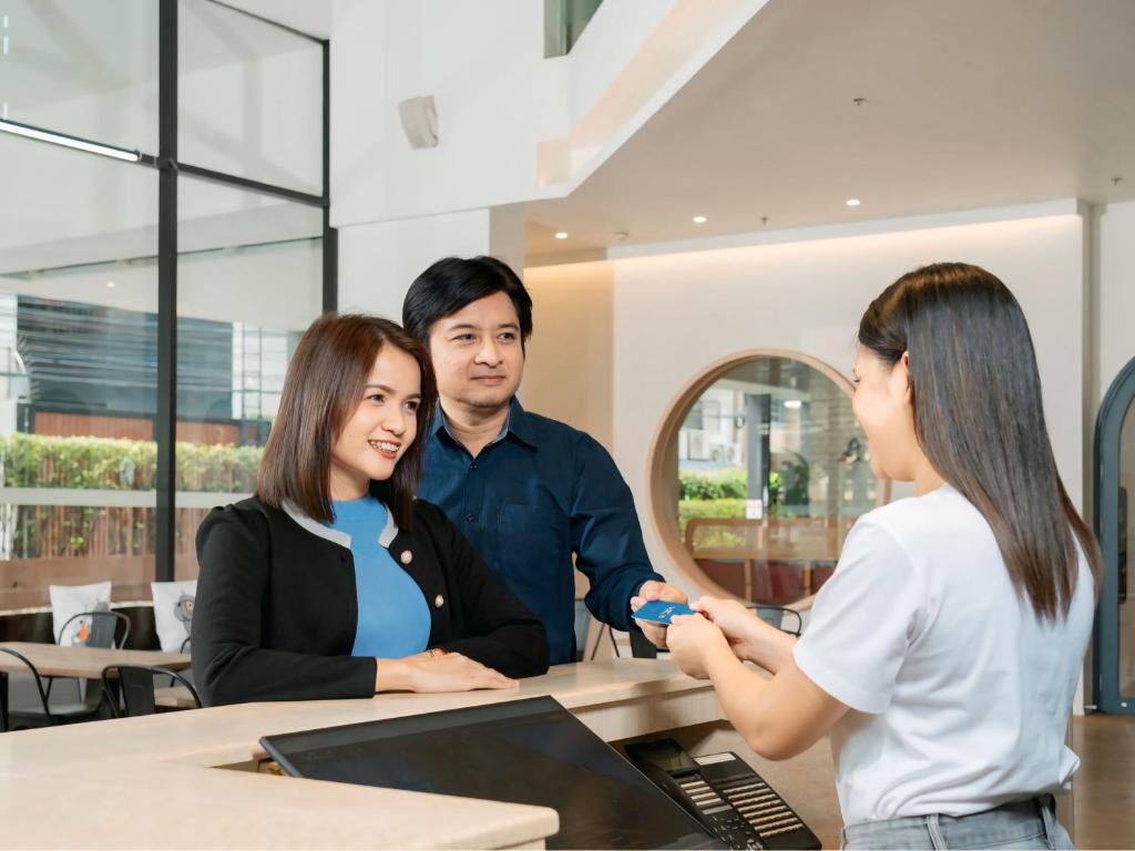 A bright hotel reception scene with a staff member assisting two guests at a curved counter.