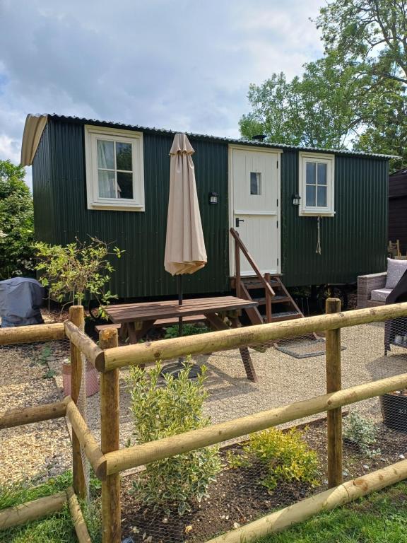 a green tiny house with an umbrella and a patio at Merrywell Shepherds Hut & Paddock in Offenham
