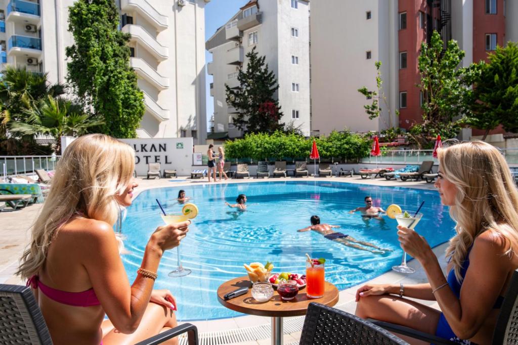 Zwei Frauen sitzen mit Getränken an einem Swimmingpool. in der Unterkunft Marmaris Alkan Hotel in Marmaris