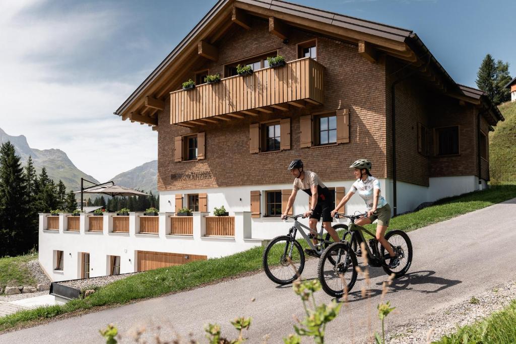 two people riding bikes in front of a house at Bergmähder - Alpine Residence in Oberlech