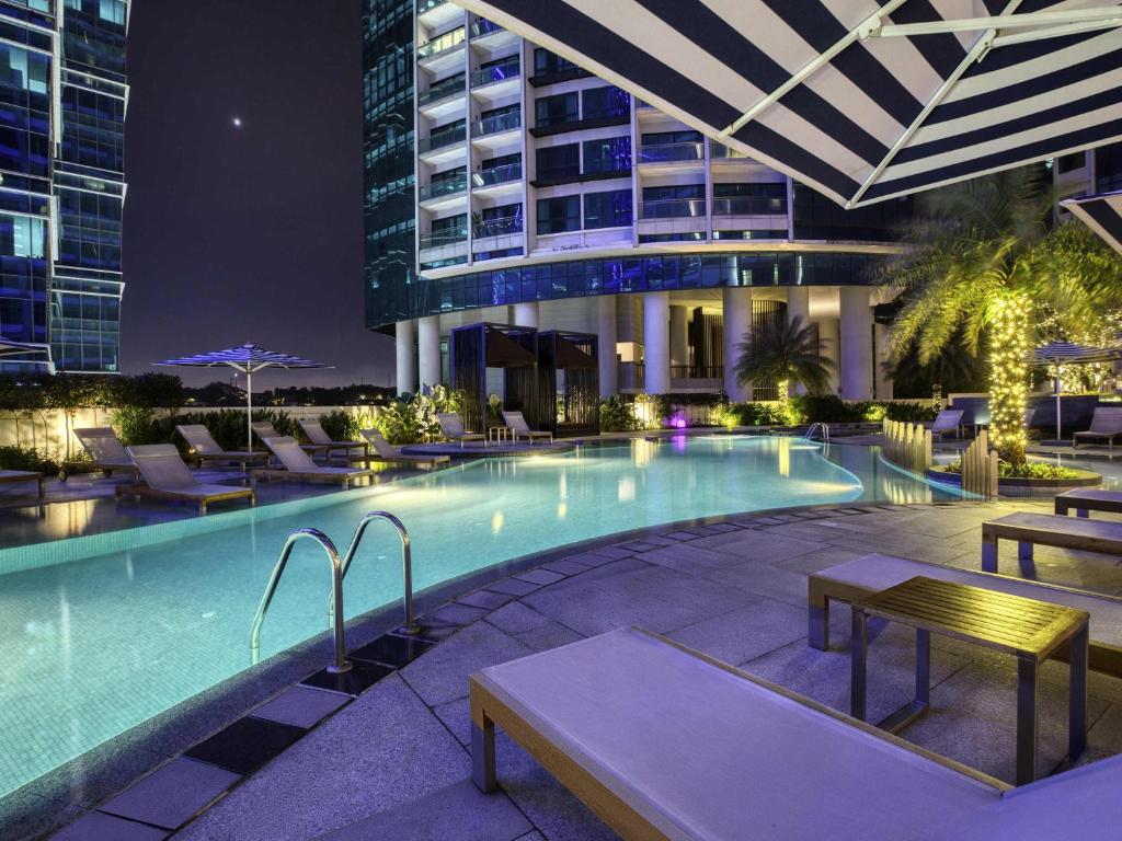 A rooftop-style pool deck lit for evening use, with loungers, tables and city buildings around the water.