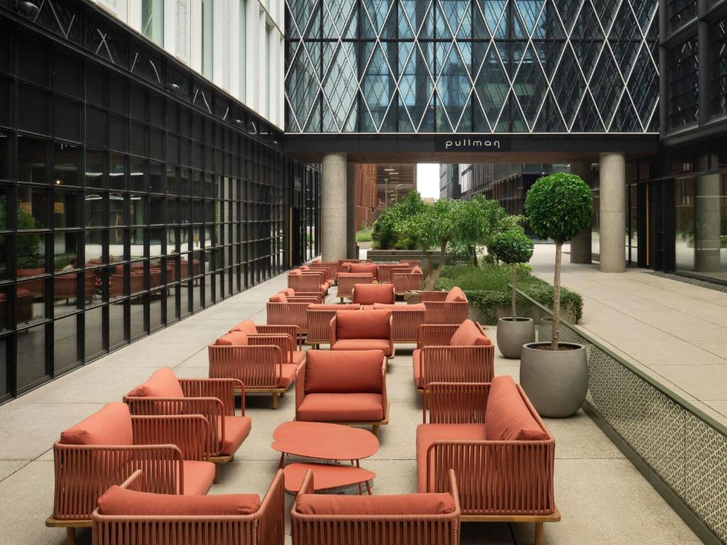 Outdoor lounge with orange woven sofas and round coffee tables in a glass-enclosed courtyard with a central tree and 'pullman' sign at Pullman Zagreb in Zagreb