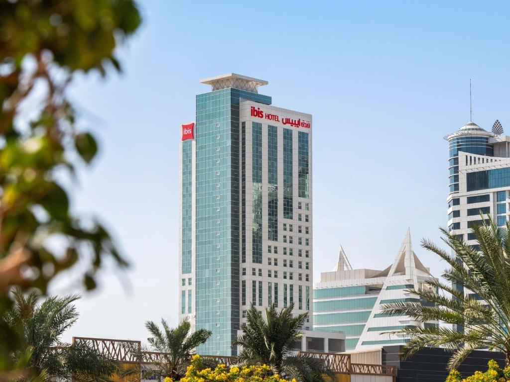 a tall glass building with a red sign on it at ibis Seef Manama in Manama