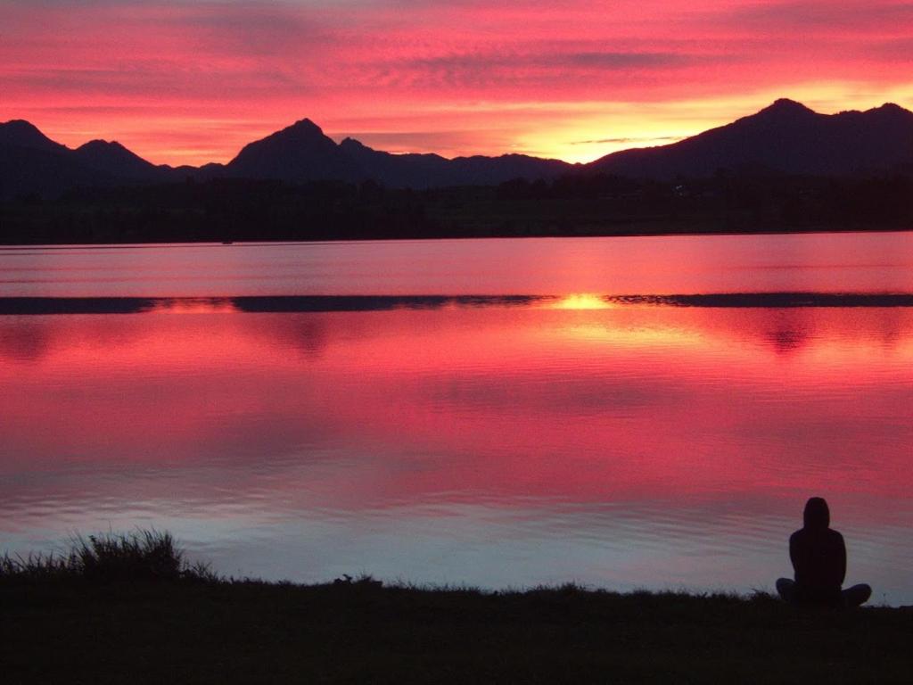a person sitting on the shore of a lake at sunset at Apartment mit Seeblick - Hopfen am See in Füssen