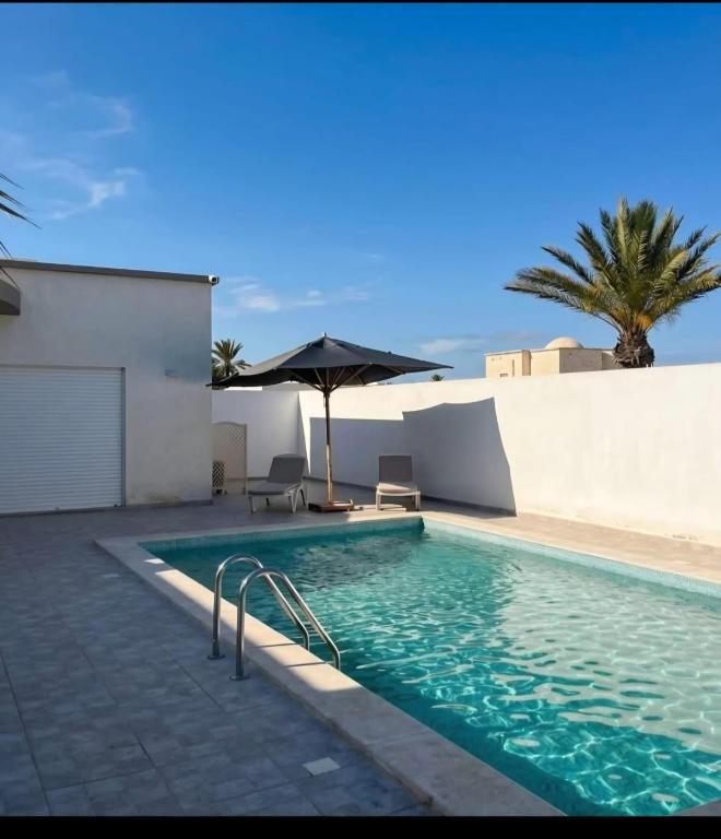 a swimming pool with an umbrella and chairs and a palm tree at Villa Wissal Djerba piscine privée in Arkou