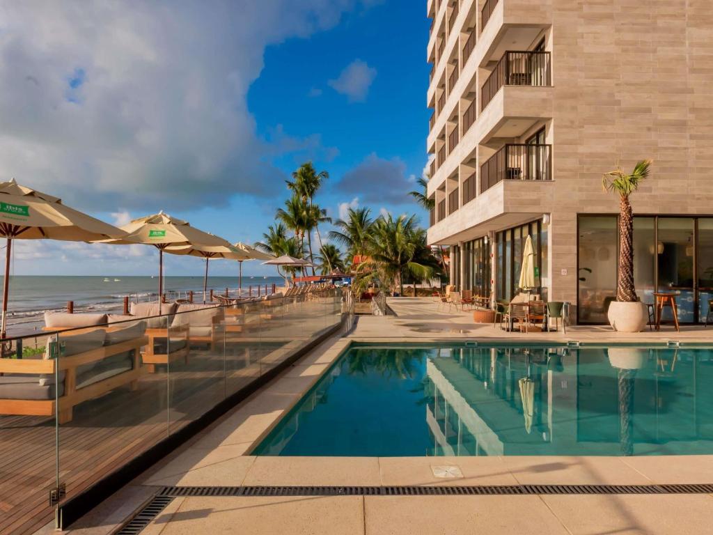 a hotel pool with chairs and umbrellas and the ocean at Ibis Styles Maragogi in Maragogi