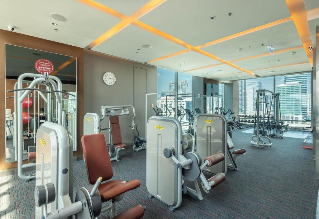 A bright fitness room contains resistance machines and cardio equipment beside city windows.