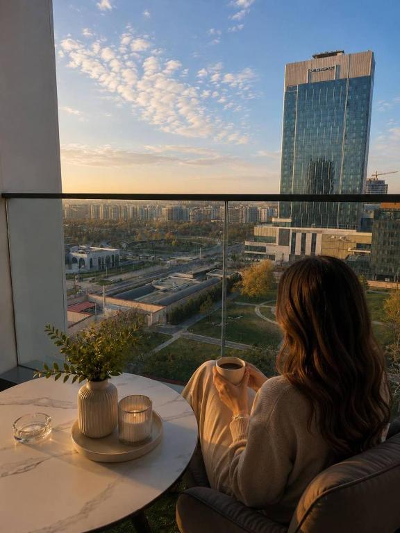 a woman sitting at a table looking out of a window at AsiaWay Apartment-NEST ONE in Tashkent