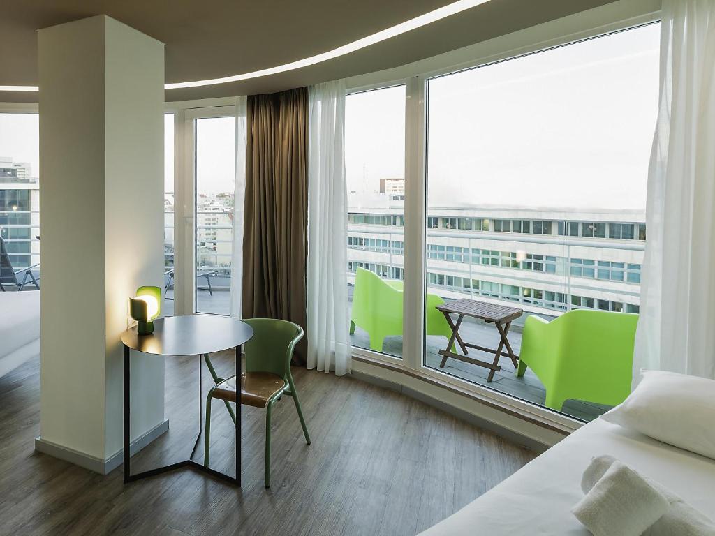 A room corner with a bed, small dining table, green chairs, and broad glazed doors opening to a balcony.