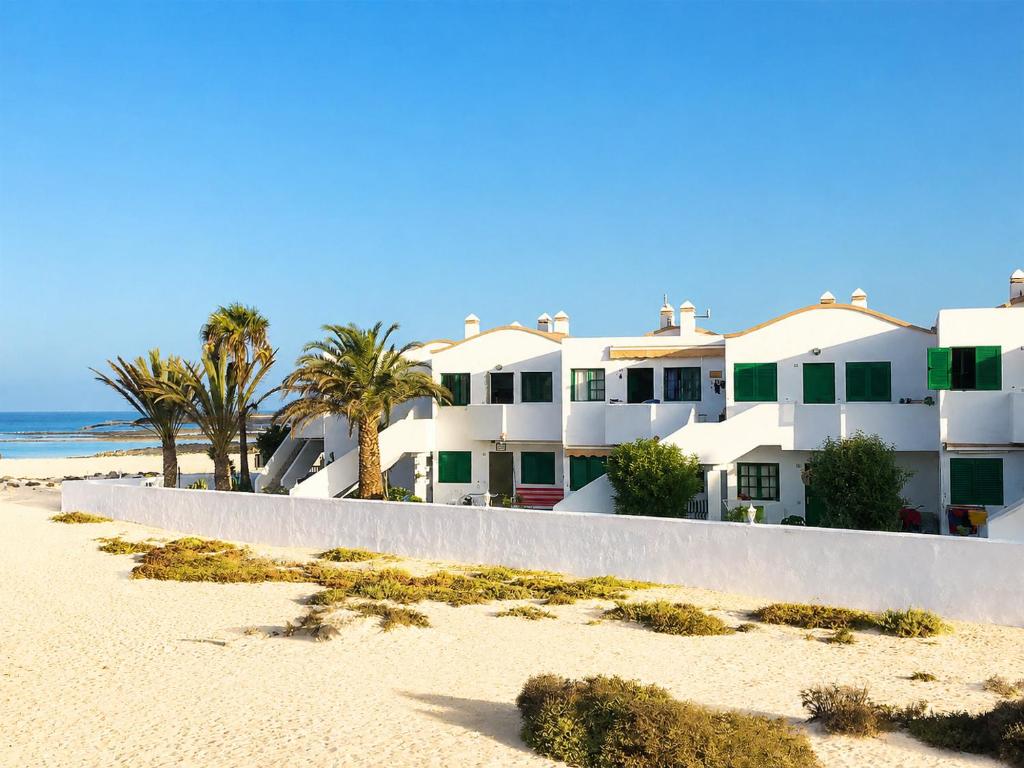 a white building on the beach with palm trees at Apartamento Los Lagos Renovated New Kitchen & More in El Cotillo