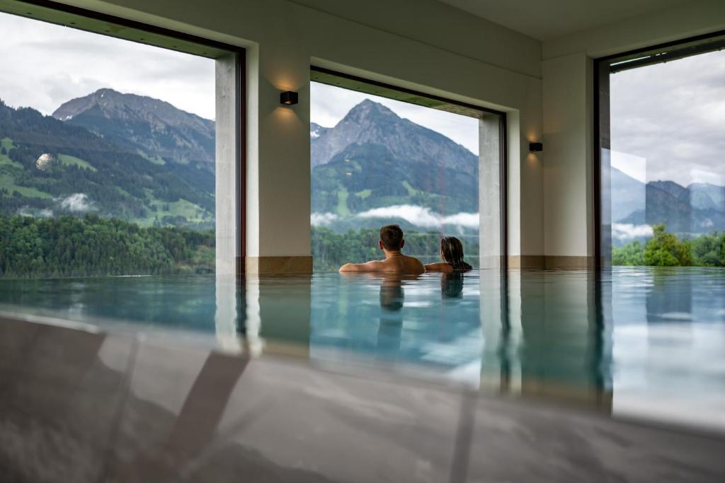 a man and a woman sitting in a swimming pool with mountains at AllgäuHotel Tanneck in Fischen