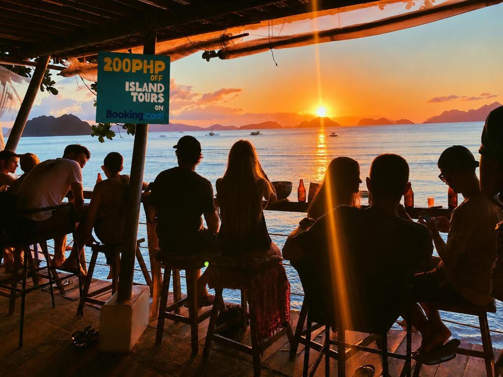 a group of people sitting at a bar watching the sunset at Outpost Beach Hostel in El Nido