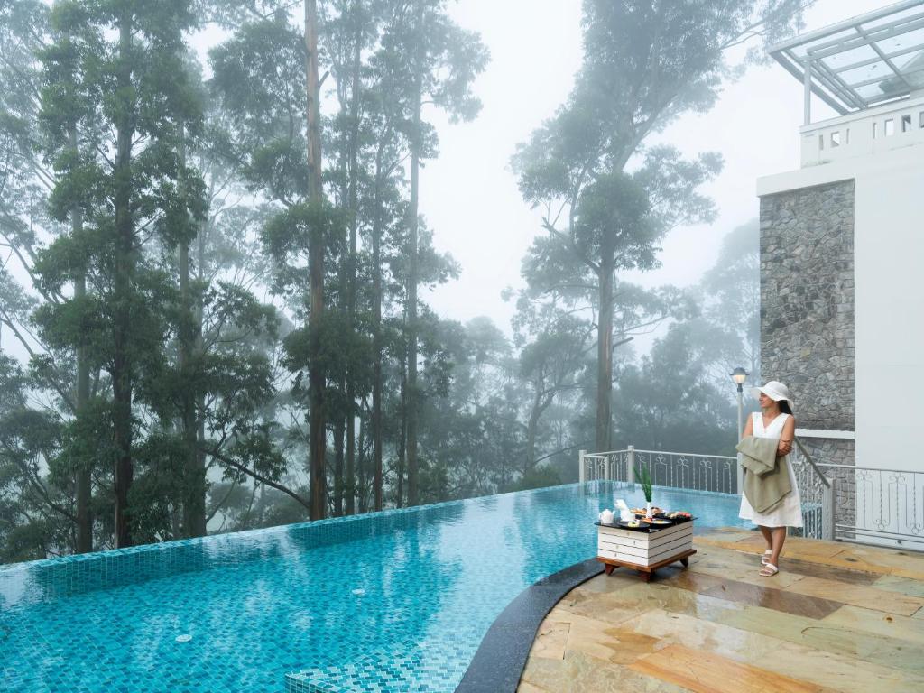a woman standing on the balcony of a resort with a pool and trees at 1924 Clock House Munnar in Munnar