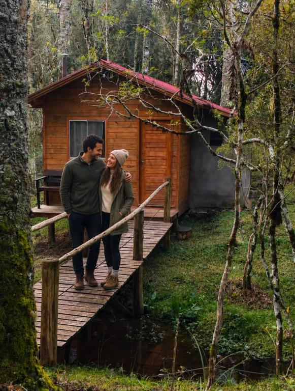 a man and woman standing on a wooden bridge in front of a cabin at Cabana Recanto dos Aparados in Cambará